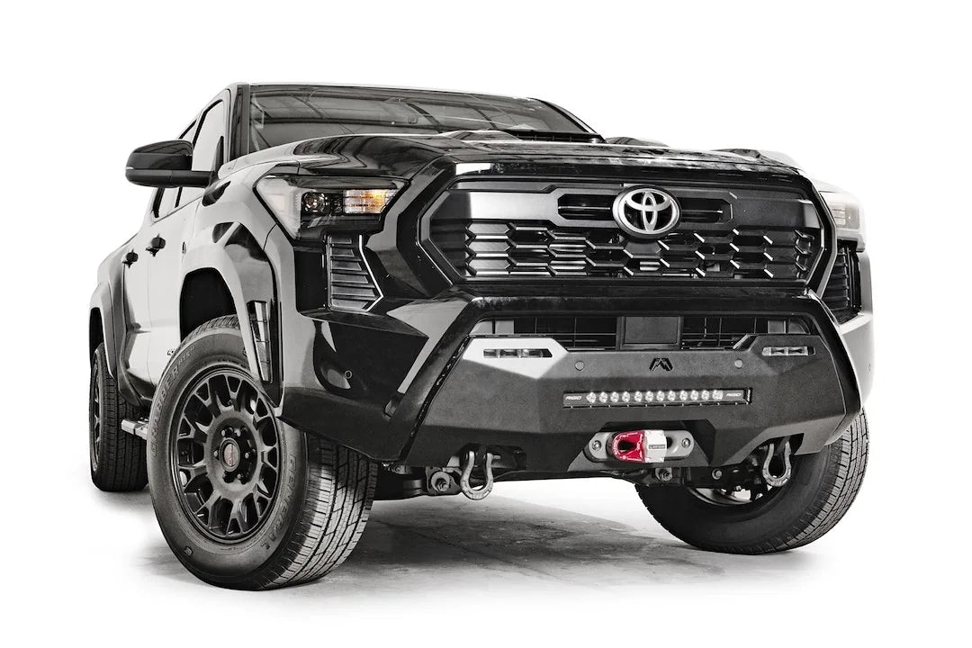 Fab Fours Hidden Winch Mount on a black Toyota Tacoma, front three-quarter view, showing the hidden winch with red hook, LED light bar, and D-ring shackles
