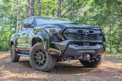 Fab Fours Hidden Winch Mount on a black Toyota Tacoma, front three-quarter view outdoors, showing the winch fairlead, LED light bar, and D-ring shackles