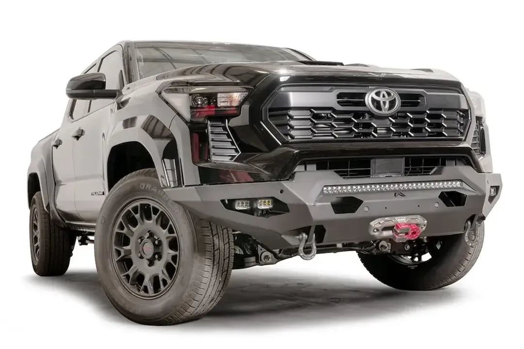 Fab Fours Matrix front bumper on a dark gray Toyota Tacoma, front three-quarter view, showing the no-guard design, LED light bar, and winch with red hook