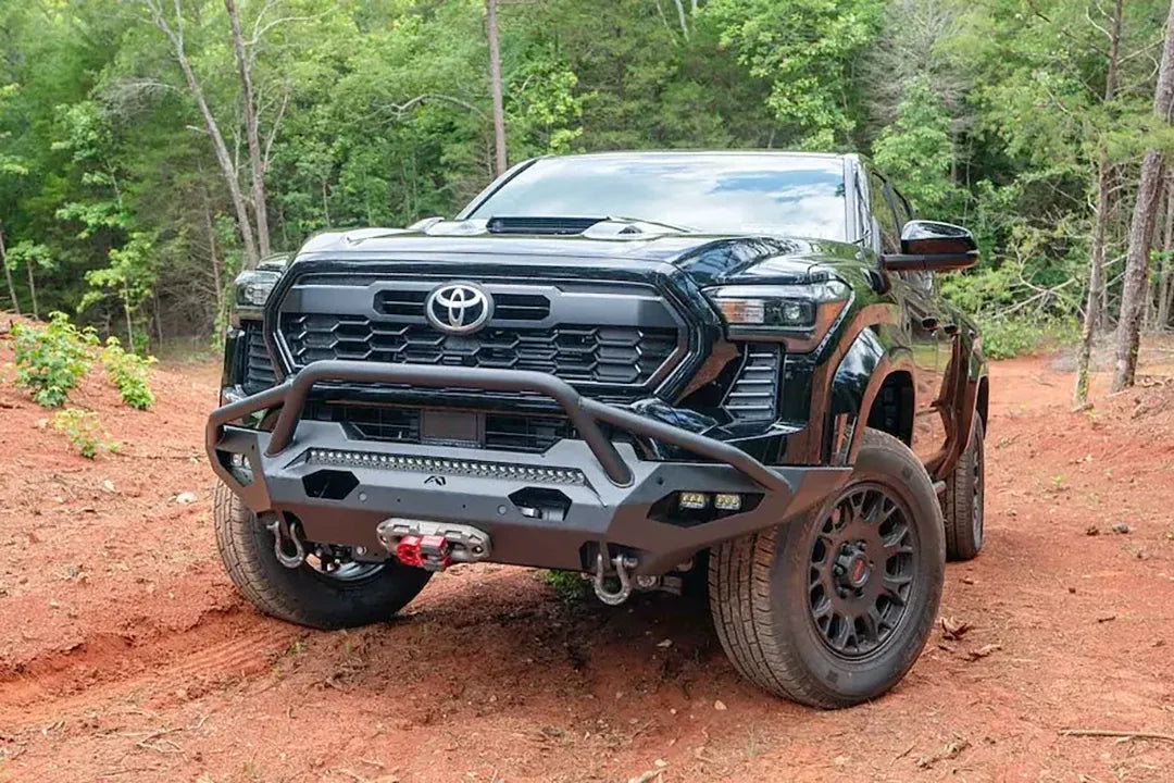 Fab Fours Matrix front bumper with pre-runner guard on a black Toyota Tacoma, front view outdoors on a dirt trail, showing the pre-runner hoop, LED light bar, and D-ring shackles