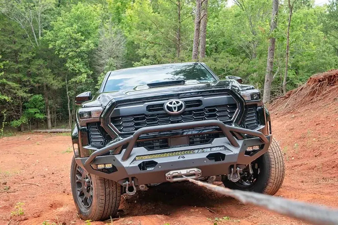 Fab Fours Matrix front bumper with pre-runner guard on a black Toyota Tacoma, front view outdoors on red clay, showing the pre-runner hoop, winch line in use, and LED light bar
