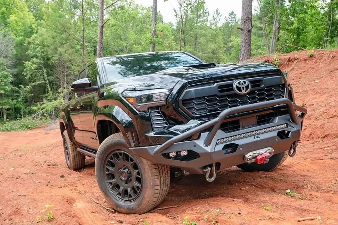 Fab Fours Matrix front bumper with pre-runner guard on a black Toyota Tacoma, front three-quarter view outdoors, showing the pre-runner hoop, cube light pods, and winch fairlead