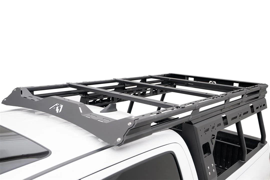 Close-up of the Fab Fours Overland Rack on a white Toyota Tacoma, showing the logo-cut wind fairing, platform crossbars and slotted side panels extending over the cab roof