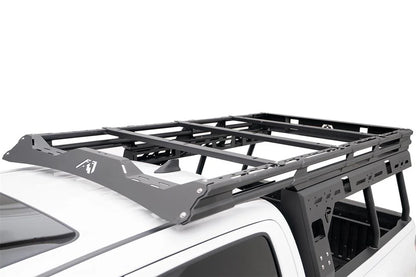 Close-up of the Fab Fours Overland Rack on a white Toyota Tacoma, showing the logo-cut wind fairing, platform crossbars and slotted side panels extending over the cab roof