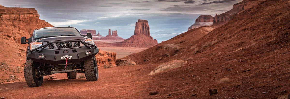 A black off-road pickup truck with a front bumper guard and large tires drives on a red dirt road through a desert canyon landscape with butte formations in the background.