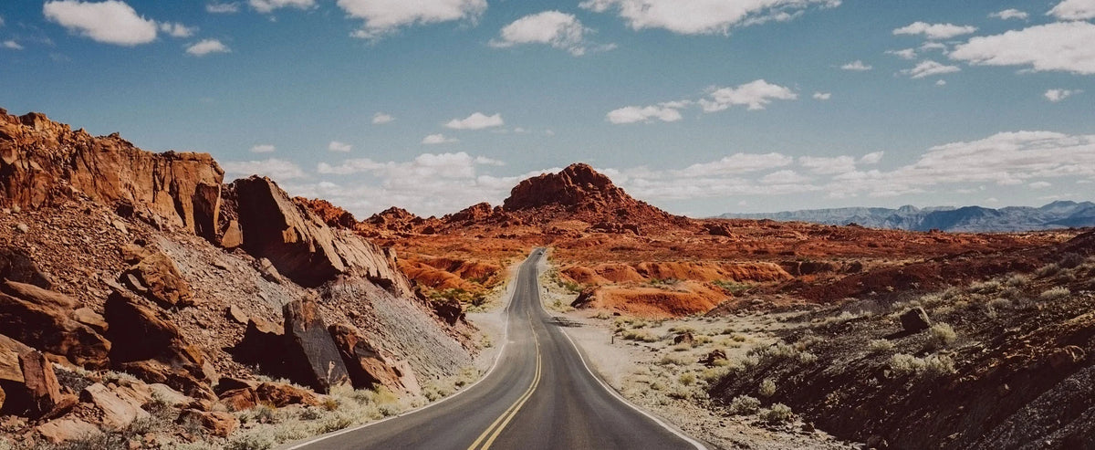 A winding paved road runs through a desert landscape with red rock formations, shrubs, distant mountains, and a blue sky with scattered clouds.